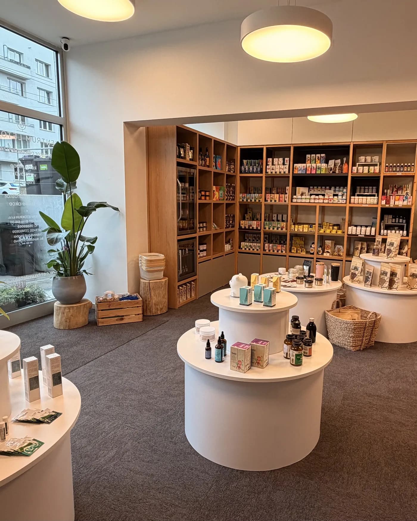 Prodejna po rekonstrukci — kulaté stolky s produkty, dřevěné regály a rostliny u okna